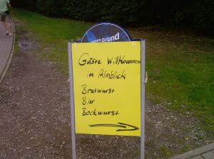 Begrüßungsschild der Gaststätte "Almblick" in Bielefeld, in schwarzer Schrift steht auf gelbem Untergrund: Gäste willkommen im Almblick, Bratwurst, Bier, Bockwurst. Darunter weist ein Pfeil nach rechts den Weg.