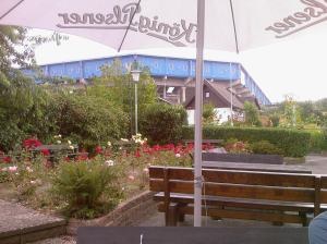 Vorn eine Holzbank unter einem weißen Sonnenschirm, links ein Beet mit blühenden Blumen, im Hintergrund ein Fußballstadion (Außenansicht)