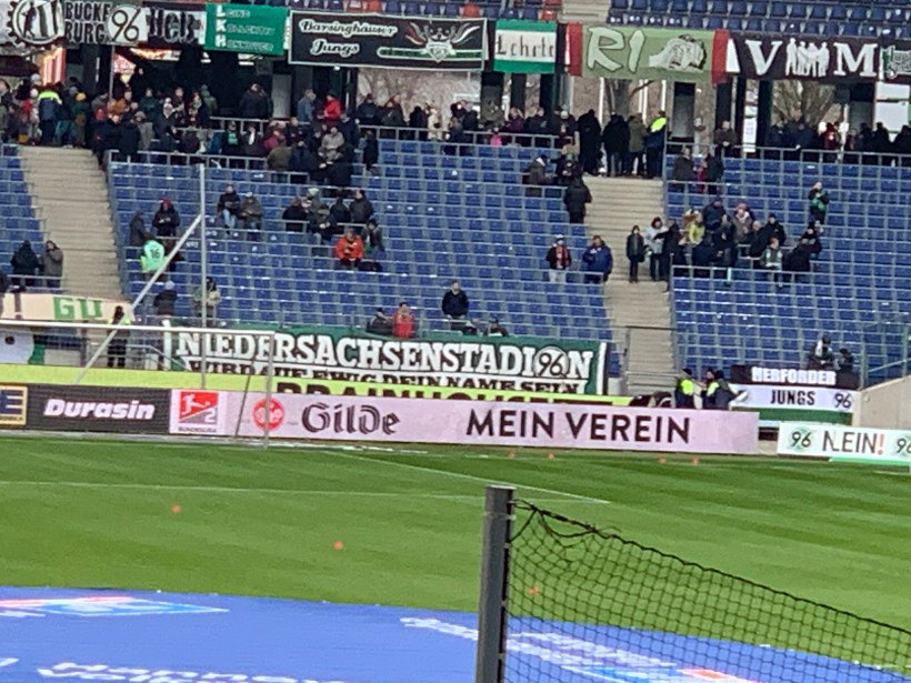 Ansicht der Nordkurve im Stadion von Hannover 96. Vor den dunkelblauen Stuhlreihen hängt eine Zaunfahne mit dem traditionellen Stadionnamen "Niedersachsenstadion