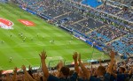 Blick vom Oberrang im Volksparkstadion auf das Spielfeld. Dieses leuchtet grün auf der rechten Bildseite, links dahinter sind die Ränge der Osttribüne zu sehen (blau)