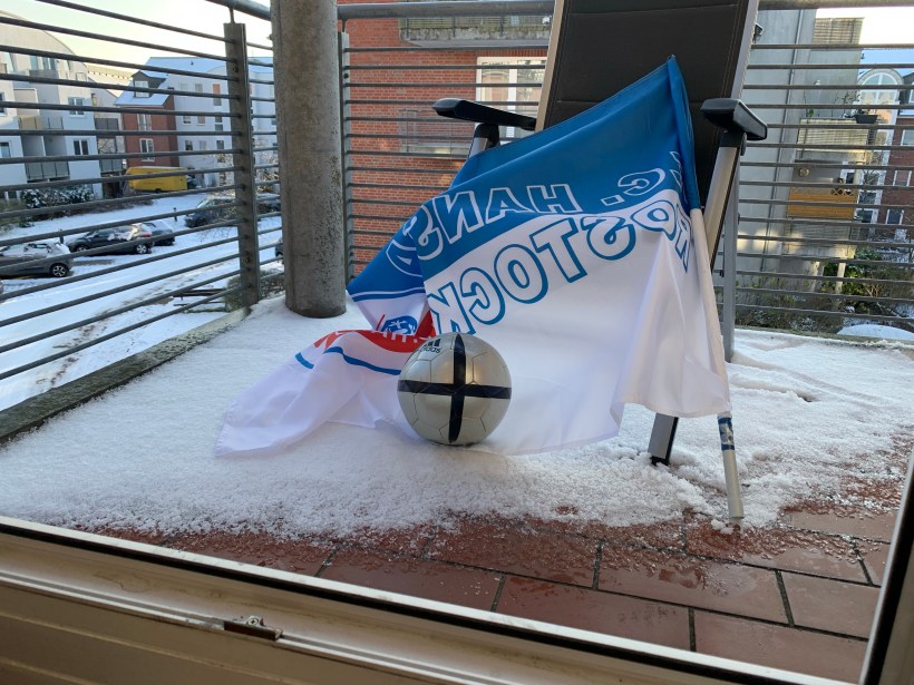 Blick durch die Balkonbrüstung in einen Innenhof. Es liegt Schnee, auch auf dem Balkon. Im Schnee ein Campingsessel, an dem eine Fahne von Hansa Rostock lehnt, davor liegt ein Fußball.