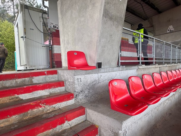 Rote Stadionsitze auf einer Betonkonstruktion mit Treppenstufen, im Hintergrund ein Sicherheitsmitarbeiter in neongelber Weste an einem Geländer.