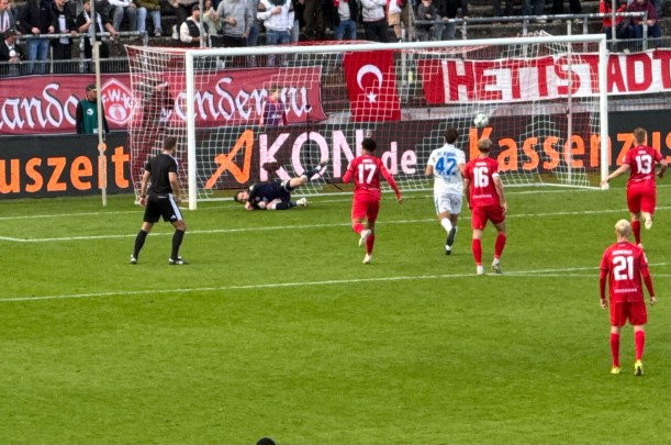 Fußballspiel auf grünem Rasen: Ein Torwart hechtet nach einem Ball, während Spieler in roten und weißen Trikots vor dem Tor auf den Ausgang warten.