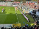 Fußballmannschaft in weiß-blauen Trikots steht auf dem Rasen vor einer vollen Tribüne, während ein Fotograf am Spielfeldrand Aufnahmen macht.