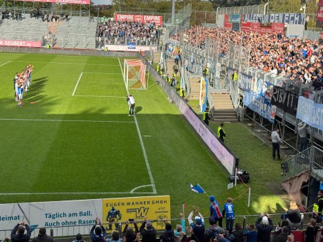 Fußballmannschaft in weiß-blauen Trikots steht auf dem Rasen vor einer vollen Tribüne, während ein Fotograf am Spielfeldrand Aufnahmen macht.
