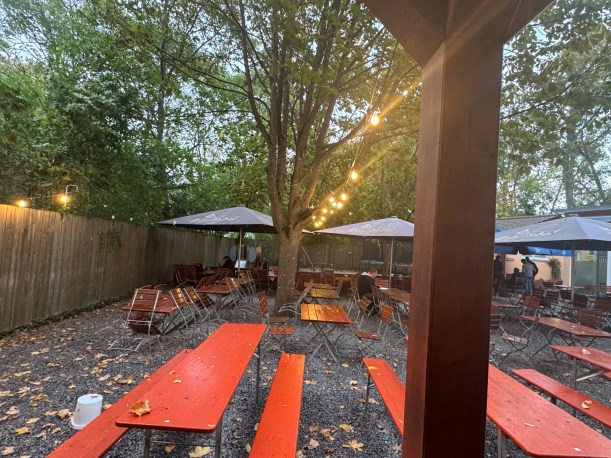 Ein Biergarten mit orangefarbenen Biertischgarnituren auf Kiesboden, umgeben von Bäumen, Holzzäunen und einer Lichterkette in der Dämmerung am Abend.